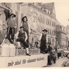 Umzug in der Stadt. Auf dem zweiten Wagen stand die badische Kuh mit ihren schwäbischen Melkern.